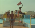 Lifeguard sitting on chair by busy pool holding up red flag Video