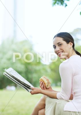 Young Woman Reading Newspaper And Eating Sandwich In City Park